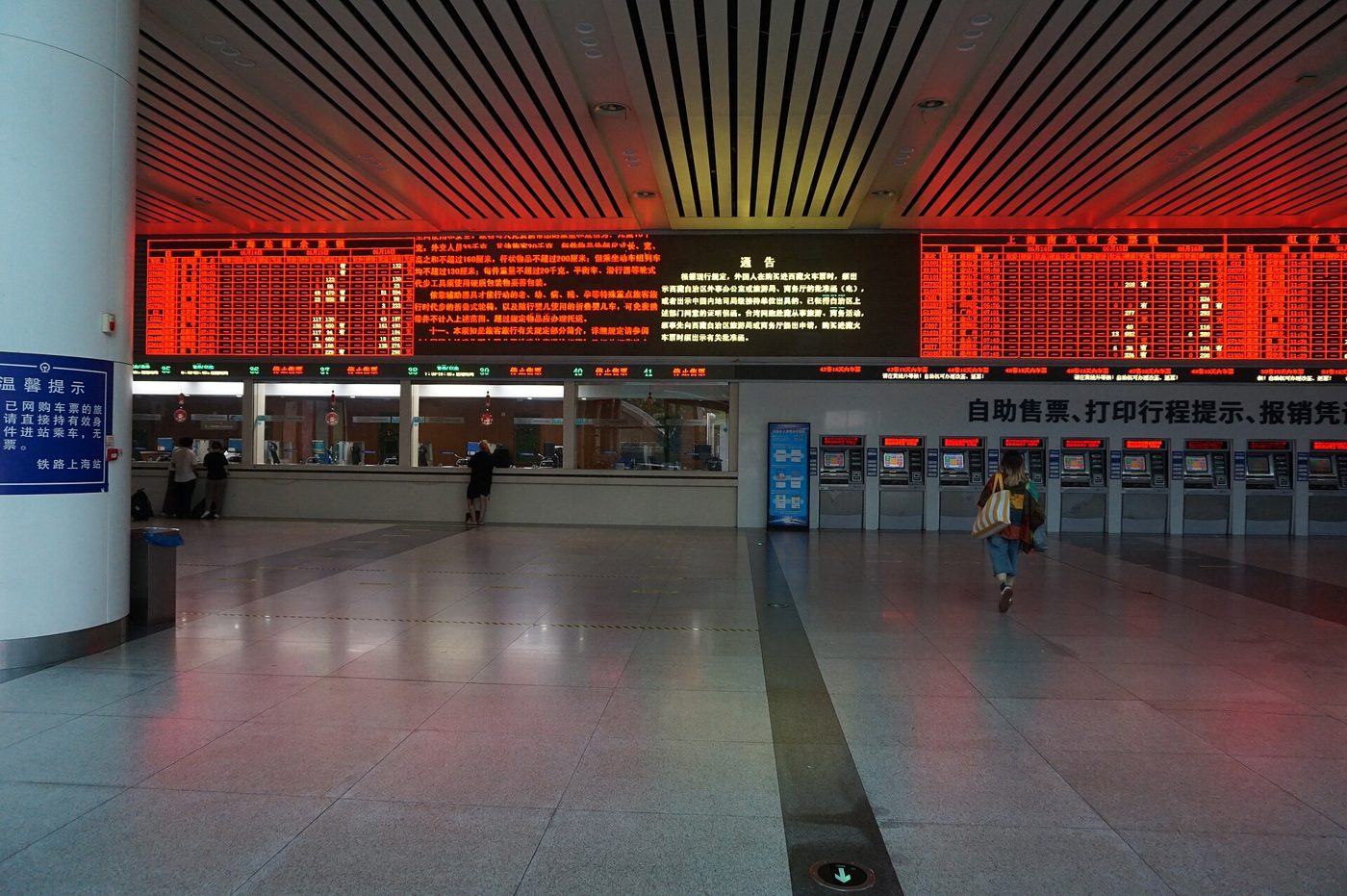A ticket hall at Shanghai Railway Station, with red departure boards overhead, staffed ticket windows on the left and a row of self-service ticket machines on the right, one traveler walking past