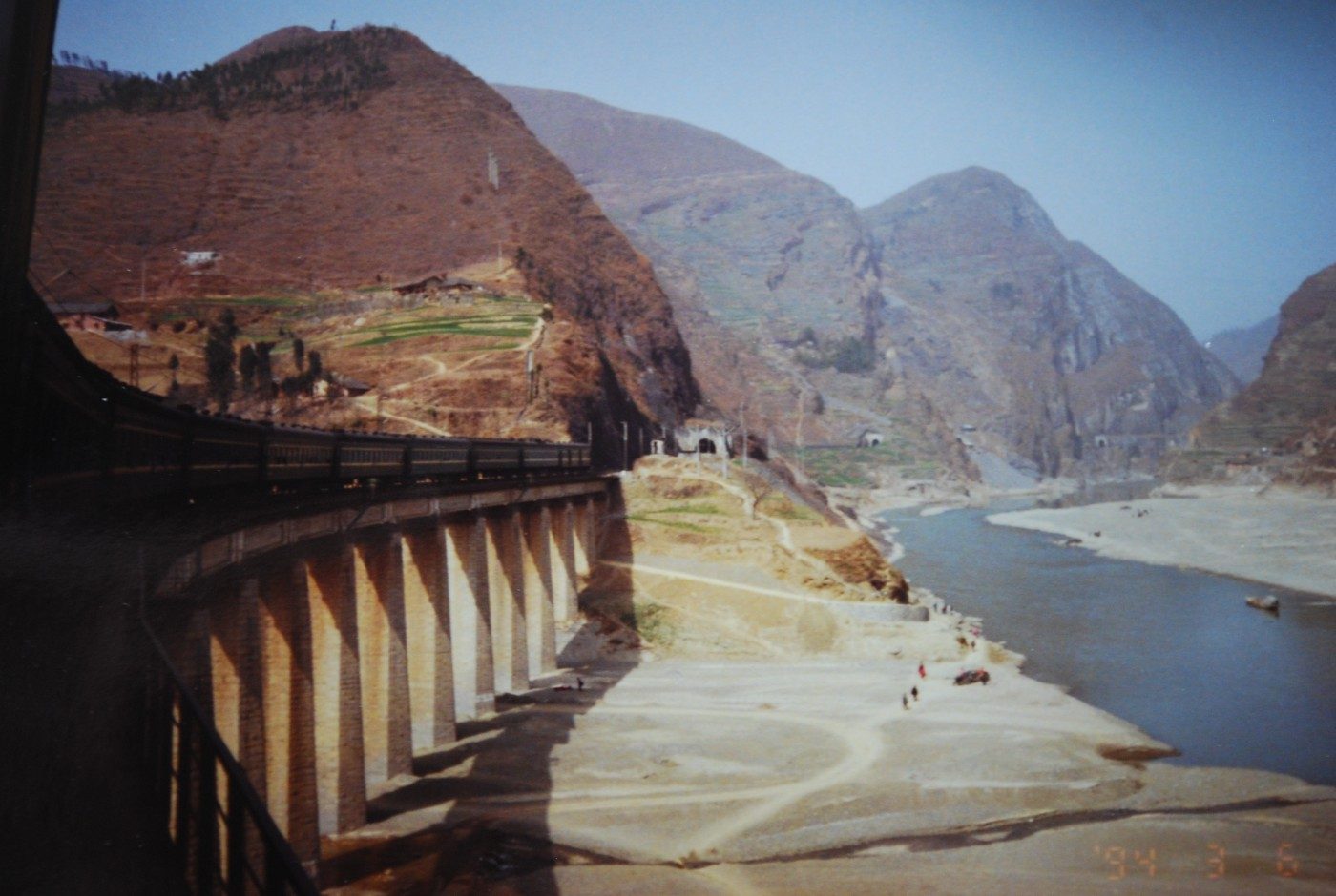 A train curving along a mountain viaduct above a dry river gorge, the kind of terrain the old Chengdu-Kunming line crosses through Sichuan's mountains.