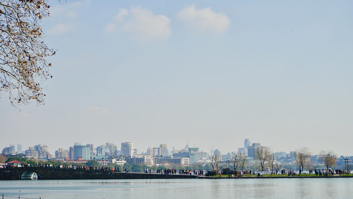 Hangzhou's city skyline seen across the calm water of West Lake, with green hills in the background.