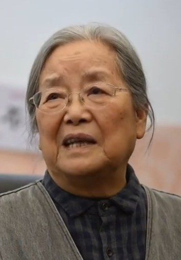 Cai Gao, an elderly Chinese woman with grey hair and glasses, wearing a grey cardigan over a plaid shirt, mid-speech during an interview