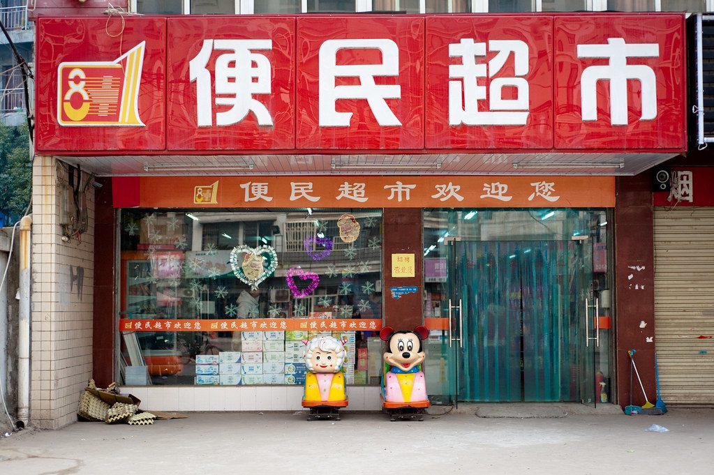 A small neighborhood supermarket storefront in Wuhan with signs advertising daily goods.