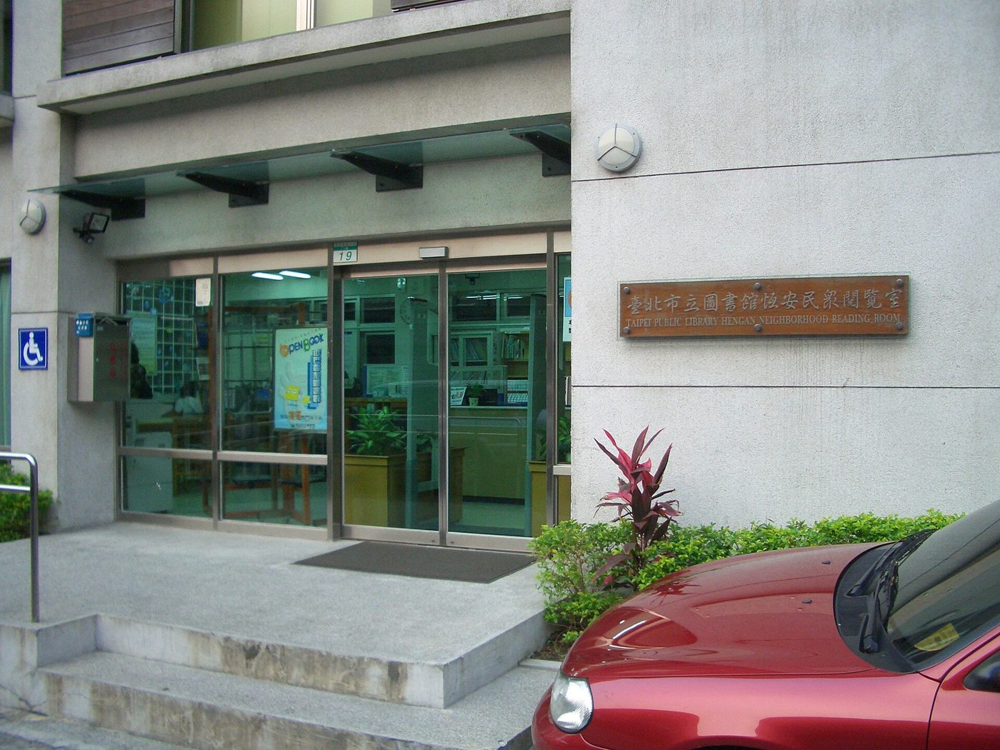 The glass sliding-door entrance of the Taipei Public Library's Heng'an Neighborhood Reading Room, with a bronze nameplate on the wall beside it and a red car parked out front.