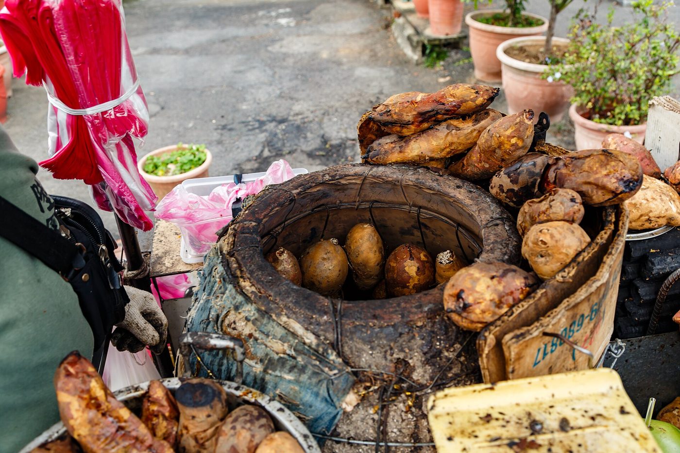 A street vendor stands beside his mobile cart piled with roasted sweet potatoes, cooking tools arranged on top, with a city street visible behind him.