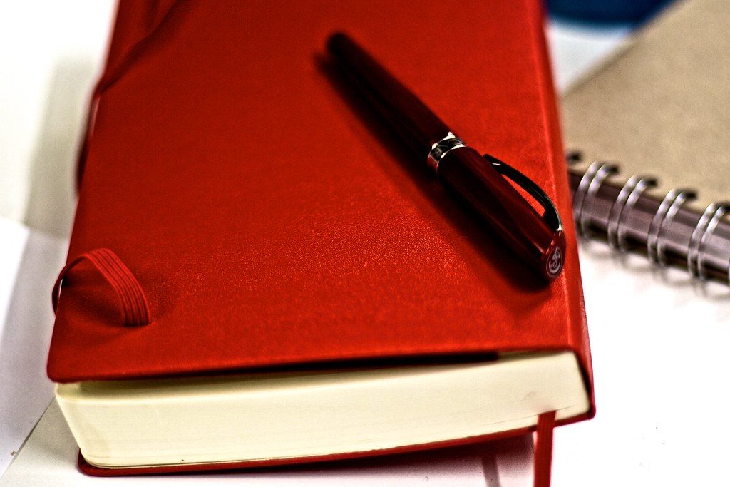 A red Moleskine diary notebook lying closed on a desk next to a pen.