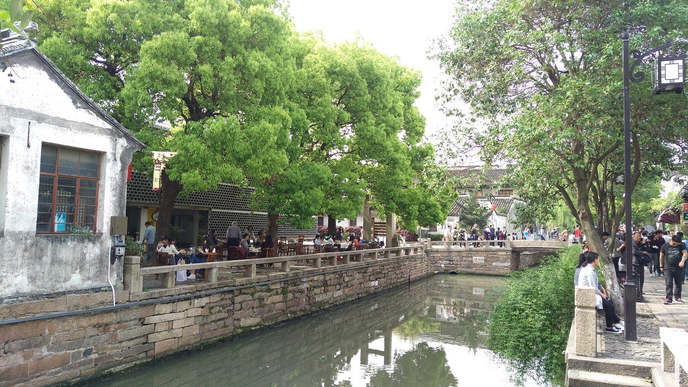 A canal in Suzhou's old Pingjiang Road neighborhood, lined with whitewashed houses, stone bridges, and green riverside plantings.