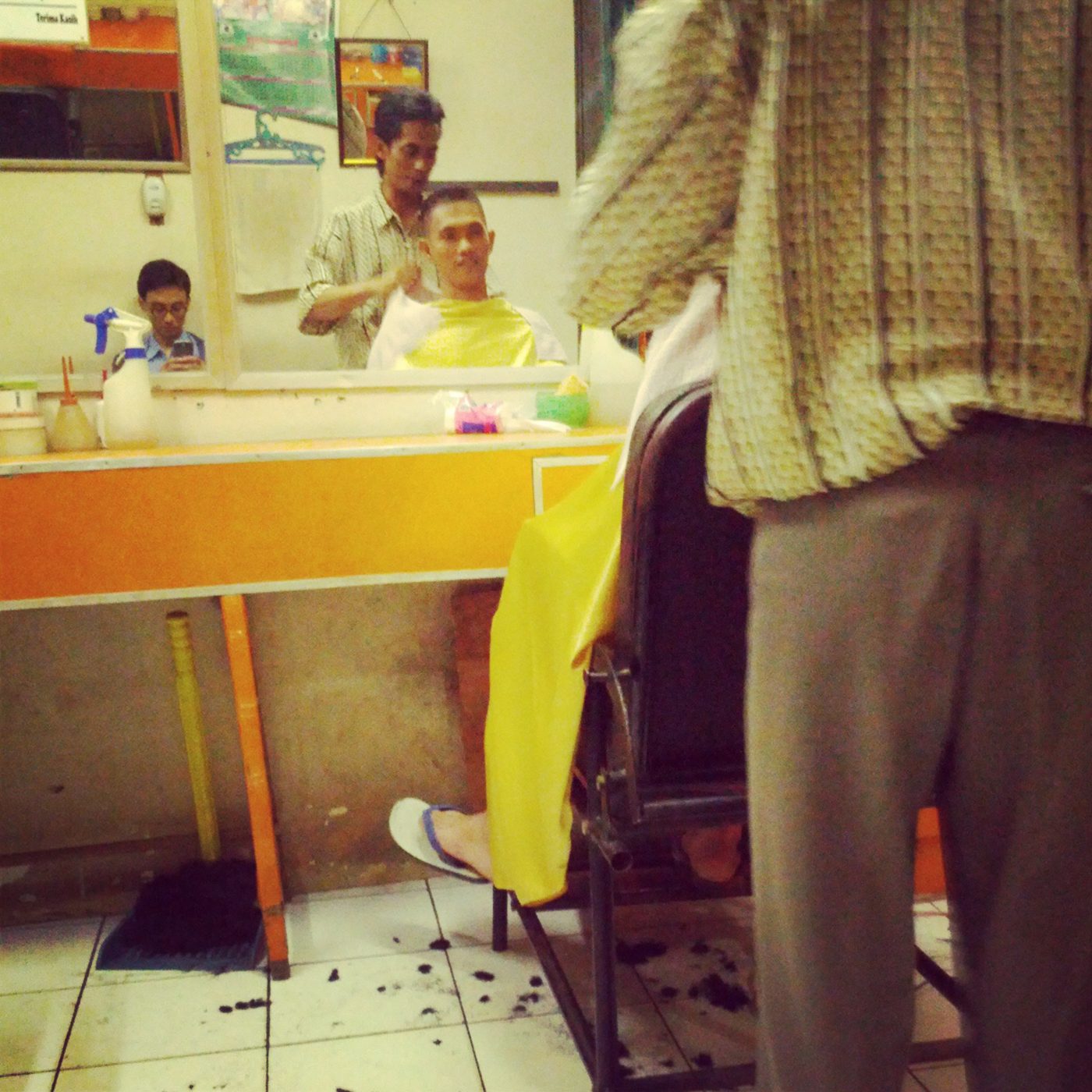 A barber in a small shop cuts a seated customer's hair in front of a mirror, with a yellow cutting cape draped over the chair and hair clippings scattered on the tiled floor.