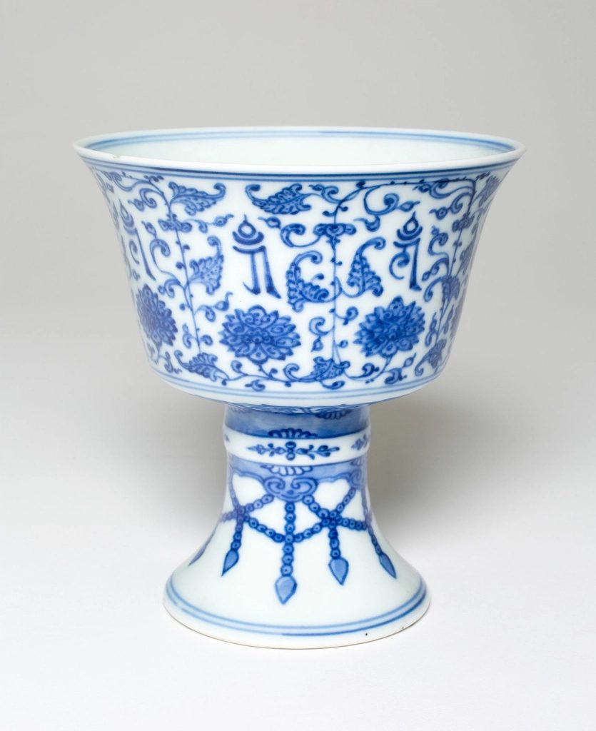 A white porcelain stem cup painted with pink peony flowers and leafy vines, with a line of decorative script near the rim.