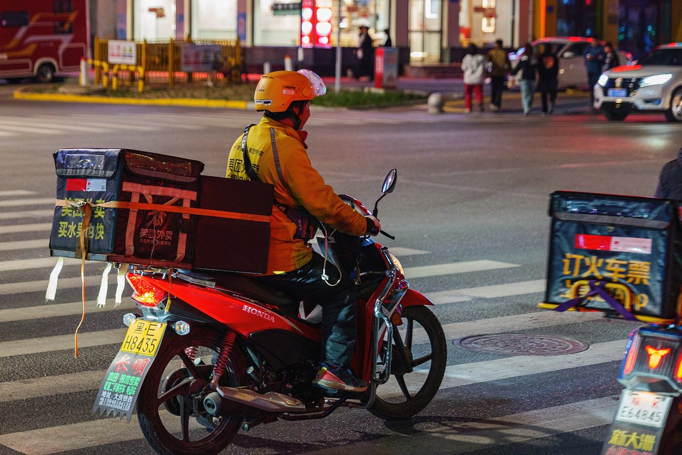A Meituan food-delivery rider in a yellow uniform waits with his scooter at a street crossing in China.