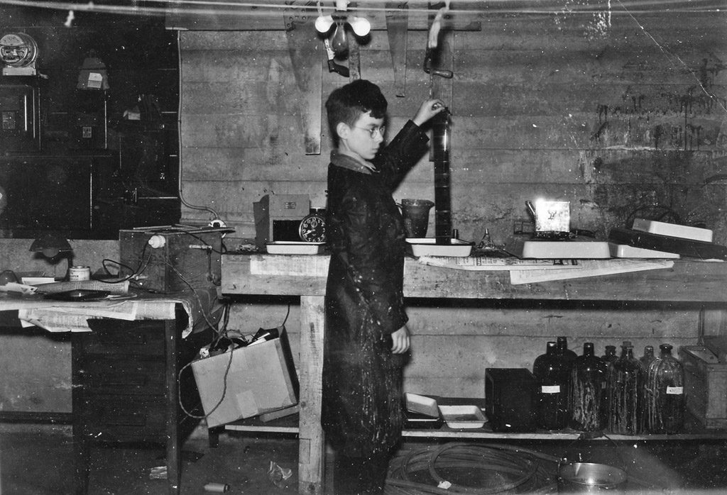 A black-and-white archival photo of a young man carefully developing film by hand under dim light in an old darkroom.