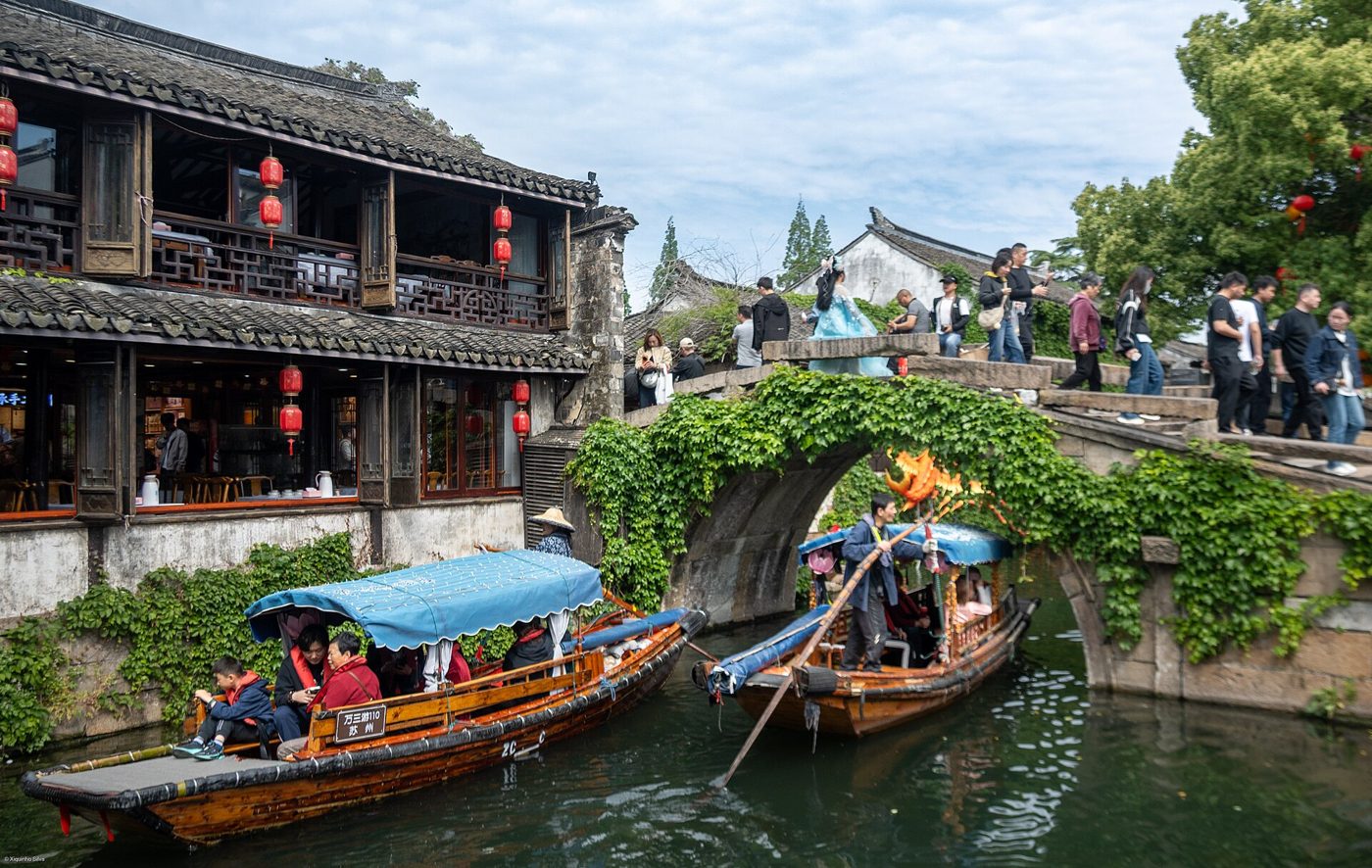 Whitewashed old houses with dark tiled roofs line a narrow canal in Zhouzhuang water town, small wooden boats moored along the water's edge.