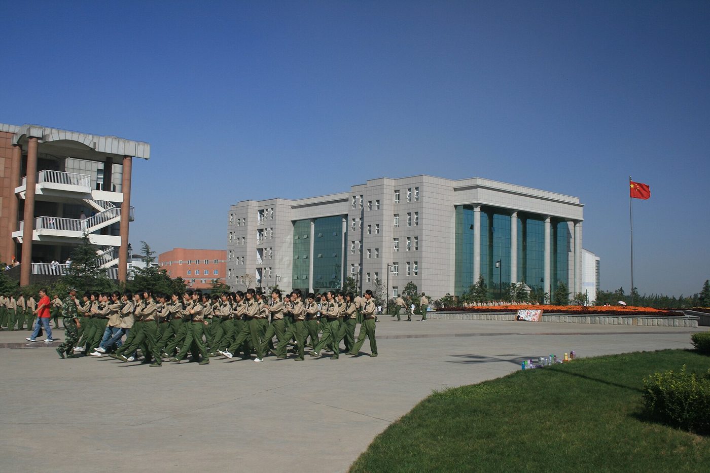 Rows of newly arrived Chinese university students in olive-green military training uniforms marching in formation across a campus square, with the national flag flying nearby