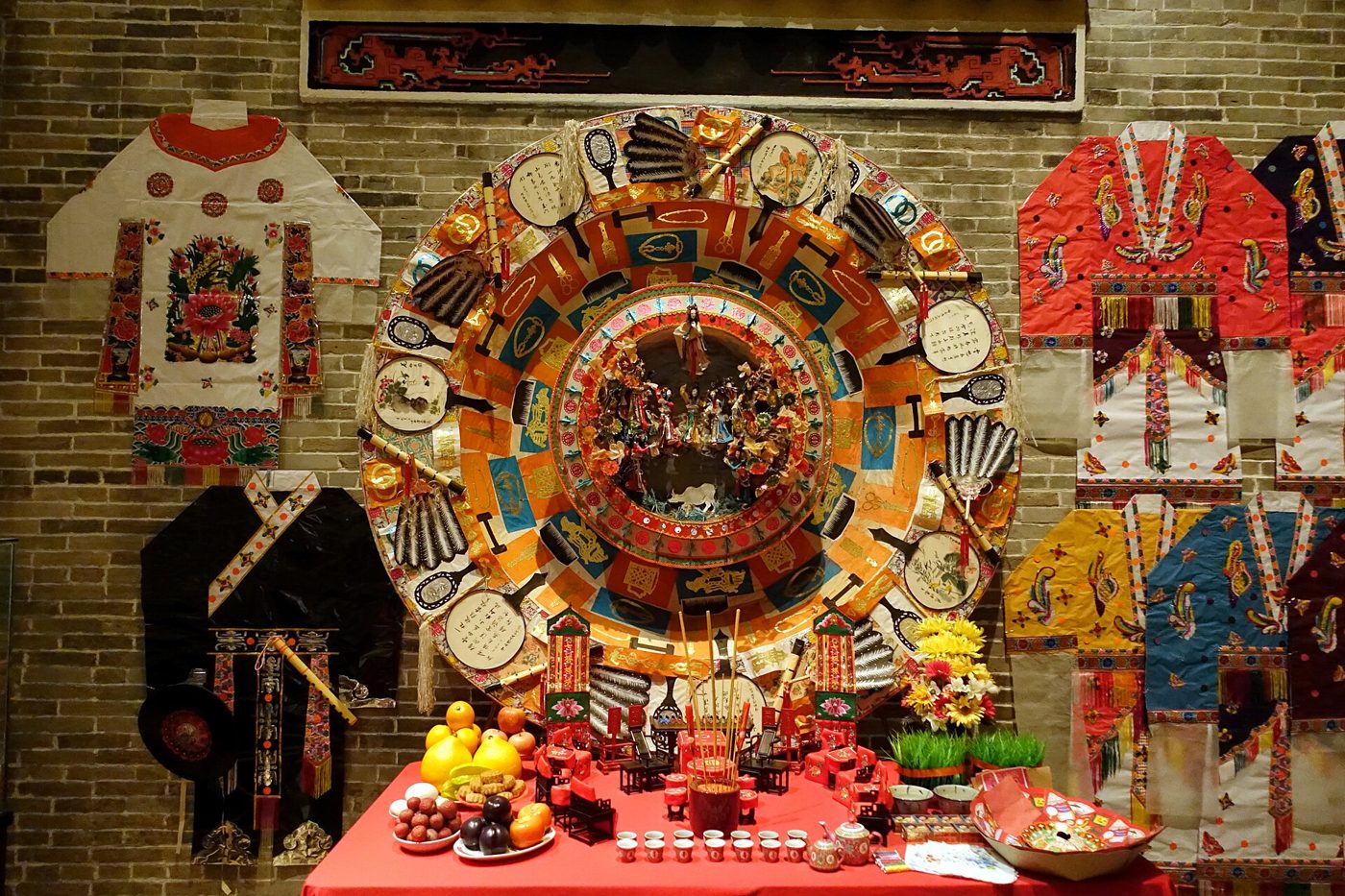 A large round Qijiepan offering wheel for the Seven Sisters festival, with tiny figures re-enacting the Cowherd and Weaver Girl legend at its centre, flanked by embroidered garments and a red altar table with fruit and incense, in the Hong Kong Museum of History.