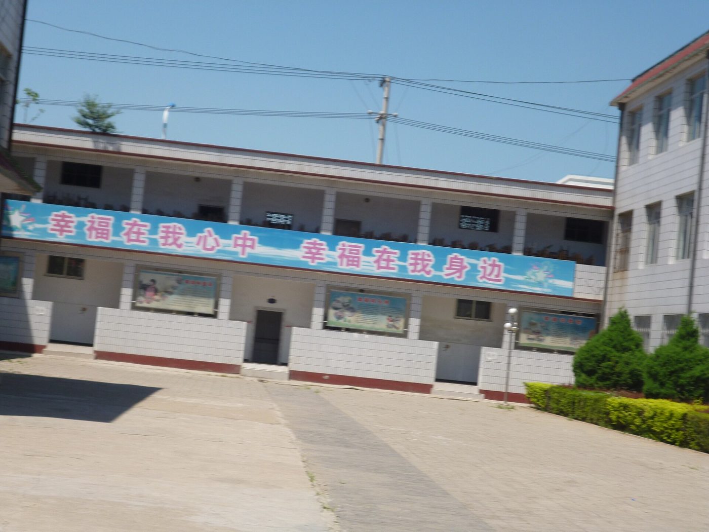 A two-storey village school in Yutian County, Hebei: a long blue banner reading 「幸福在我心中 幸福在我身边」 runs along the front wall above glazed notice boards, and the concrete yard in front of it is empty.