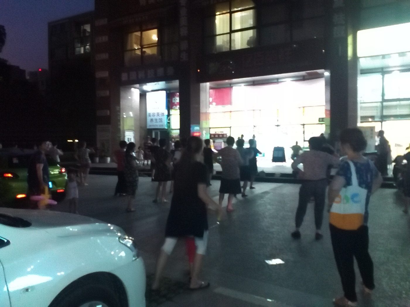 Neighbors dancing in an open lot at night outside a brightly lit shopfront, with parked cars in the foreground and lit apartment windows above.
