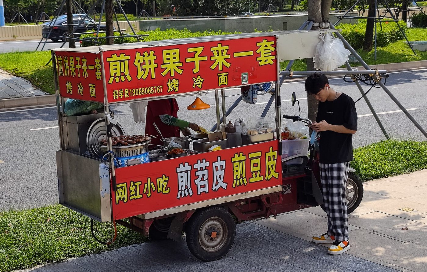 A street food vendor's cart in China with a young man standing beside it, waiting for his snack.