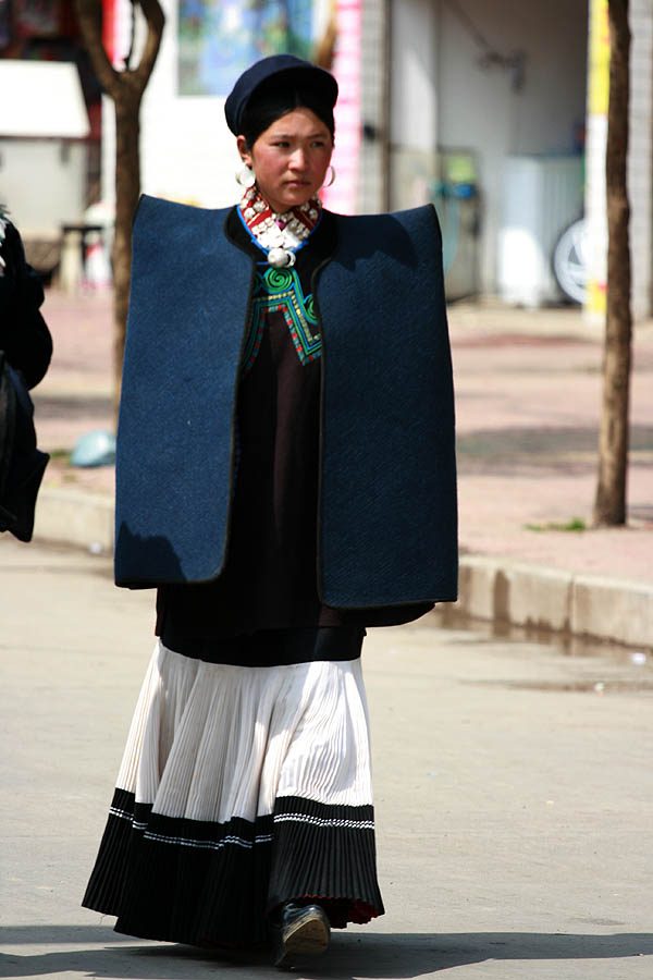 A Yi woman in a traditional dark blue felt cape and pleated black-and-white skirt walking down a street in Butuo County, Sichuan
