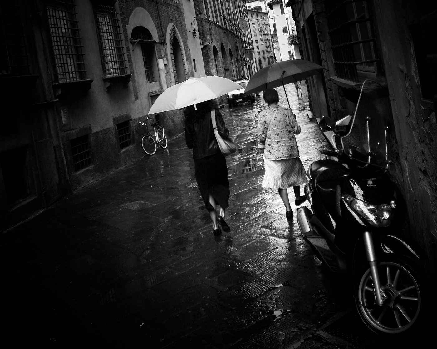 Two people walk with umbrellas down a narrow rain-wet street in Siena, Italy, a bicycle parked against the wall beside them.