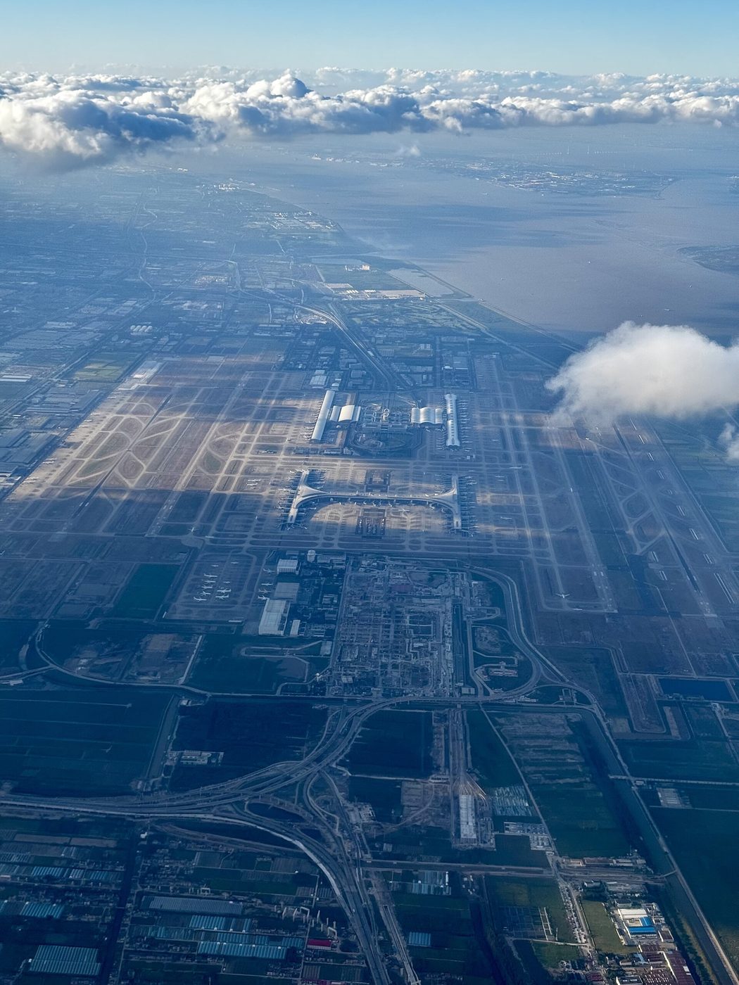 Aerial view of Shanghai Pudong International Airport's terminals and runways beside the coast, with clouds building overhead
