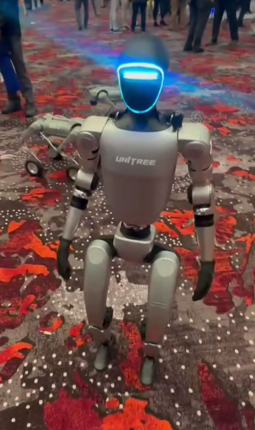 A Unitree G1 humanoid robot walking through a crowd at a trade expo, its screen-like face lit blue