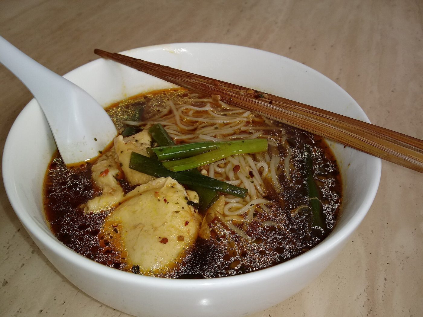 A white bowl of noodle soup with dark spiced broth, chicken pieces, noodles, and scallions, with chopsticks and a white spoon resting across the bowl on a wooden table.