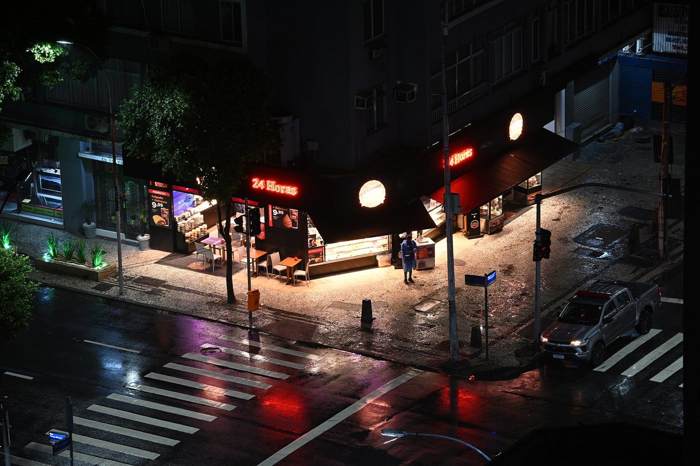 A rain-soaked city street corner at night, shopfront lights glowing and reflecting off the wet pavement.
