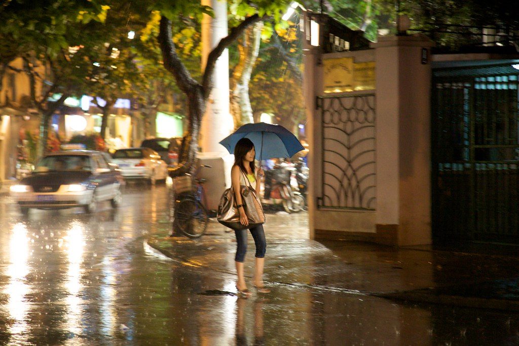 A young woman holds a blue umbrella on a rain-soaked city street at night, car headlights reflecting on the wet pavement beside a bicycle and a gated building entrance.